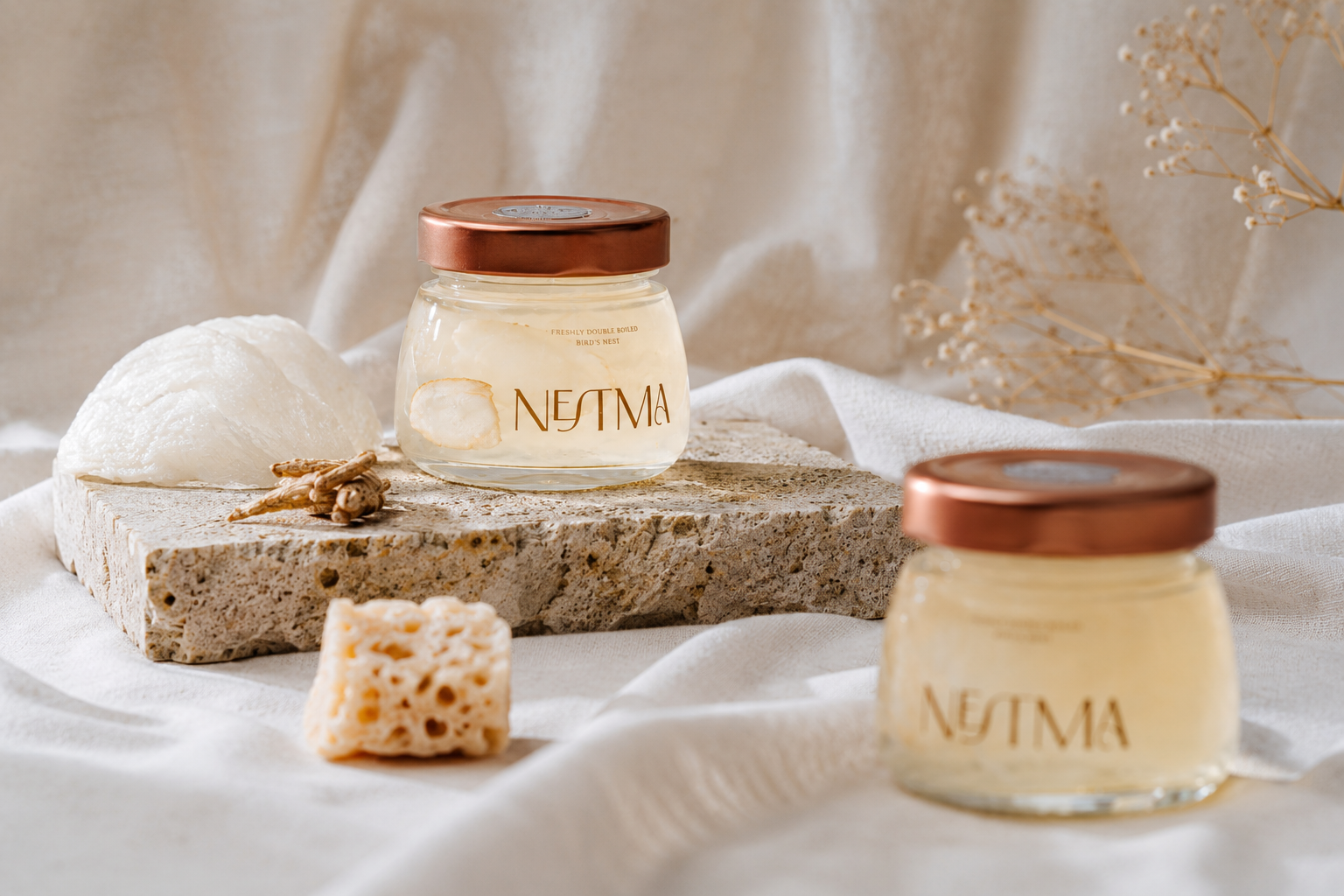 Two glass jars of NETMA premium bird's nest, styled on a stone slab with an organic raw honeycomb, and dried flowers on a draped white fabric background.